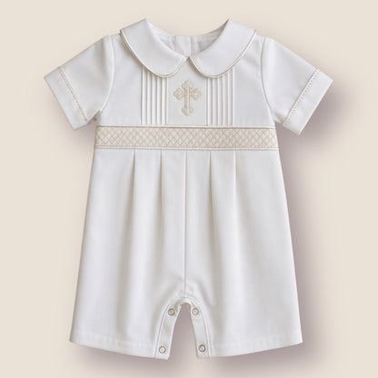 A baby boy's short-sleeved white romper with a Peter Pan collar, featuring a decorative beige-patterned band across the chest with a small embroidered cross, set against a solid beige background.